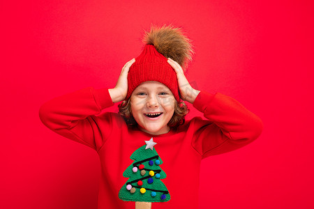 圣诞毛衣摄影照片_穿着红色圣诞毛衣的酷少年在红墙、温暖的帽子和带圣诞树的毛衣的背景下闲逛