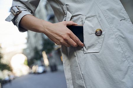 红包布口袋摄影照片_一位身穿灰色外套的女士站在户外将智能手机放入口袋的裁剪镜头