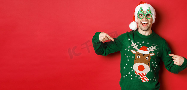 戴着派对眼镜和圣诞老人​​帽子的快乐男人的形象，指着他的圣诞毛衣，微笑着，站在红色背景上