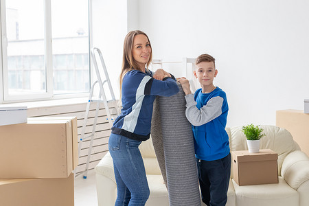 搬迁、乔迁和房地产概念 — 微笑的单亲母亲和迷人的儿子在新客厅里拿着折叠地毯，打算将其铺在新公寓里