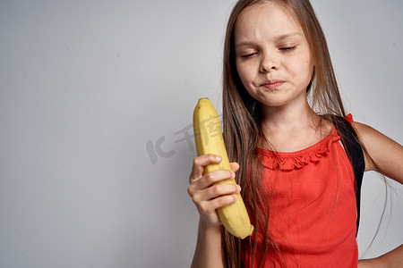 连衣裙戴眼镜背包摄影照片_戴眼镜的小女孩，穿着红色连衣裙，手里拿着香蕉，浅色背景