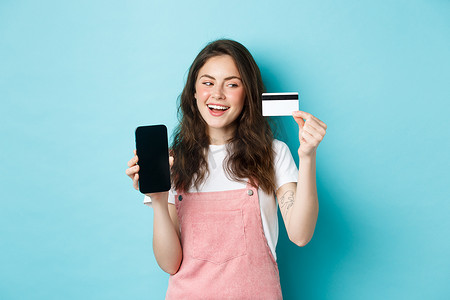 购物app摄影照片_穿着夏装的快乐女孩展示智能手机屏幕和塑料信用卡，在线支付，购物，站在蓝色背景上
