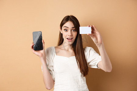 兴奋的女孩展示银行塑料信用卡和空手机屏幕，演示购物应用程序，站在米色背景上