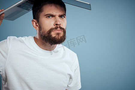 情绪化的男人，头顶上有一台打开的笔记本电脑，蓝色背景，穿着白色 T 恤，裁剪视图