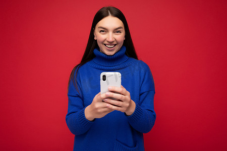 微笑迷人、积极、漂亮的年轻黑发女性，穿着时尚的蓝色温暖毛衣，在红色背景中与世隔绝，手握空白空间，使用手机短信看着相机