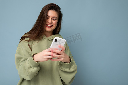 身穿时尚绿色连帽衫、微笑的美丽黑发女青年使用手机写短信，在背景中隔离，看着智能手机屏幕
