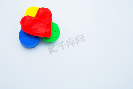 儿童游戏黄色摄影照片_白色背景上的黄色、绿色和蓝色杯子上有一颗用红色面团制成的心形模型。