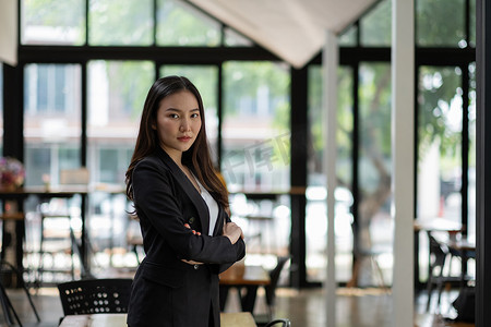 黑色西装女摄影照片_成功的年轻亚洲女商人在办公室的肖像，她站着看着镜头，穿着黑色西装。