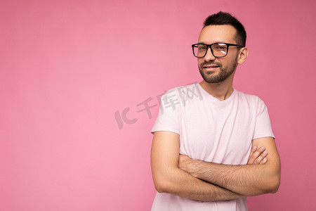 做模型摄影照片_英俊的年轻、快乐、黑发、胡子拉碴的男人，穿着白色 T 恤做模型，戴着时尚的光学眼镜，隔离在粉红色背景墙上，看着相机