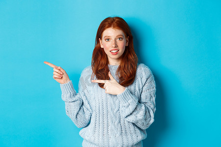 身穿毛衣的漂亮红发女孩的照片，手指指向标志，好奇地微笑着，站在蓝色背景上