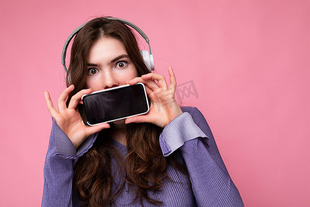 照片中，年轻的黑发卷发女性相当积极，身穿紫色短上衣，与粉色背景墙隔离，手持显示屏空空如也，蓝牙耳机看着相机的手机