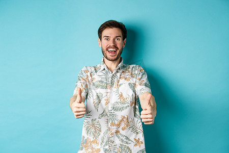 穿着夏威夷衬衫的兴奋的微笑男子竖起大拇指，看上去很惊讶，查看酷炫的促销片，站在蓝色背景上