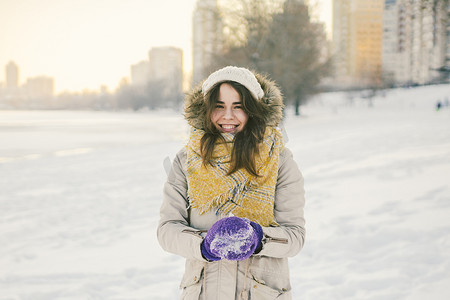一位戴着针织帽子和围巾的美丽年轻白人女子的肖像，站在冬季背景上，带着雪微笑和幸福的紫色手套，雕刻着一个雪球