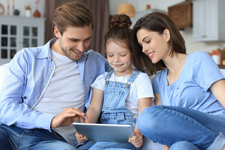 沙发椅样品摄影照片_积极友好的年轻父母与微笑的小女儿坐在沙发上一起接听数字平板电脑上的视频通话，同时周末在家放松。
