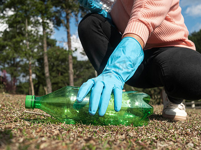 戴着手套的志愿者的近手从公园的草地上捡起一个塑料瓶。