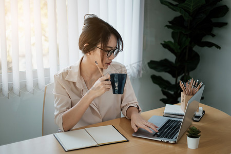 美丽的会计师坐在办公桌前的肖像，室内喝着热饮料，拿着咖啡杯，在视频会议时看着笔记本电脑。
