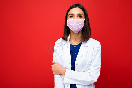 年轻漂亮的女医生脸上戴着可重复使用的病毒防护面罩，抵御红色背景墙上隔离的冠状病毒