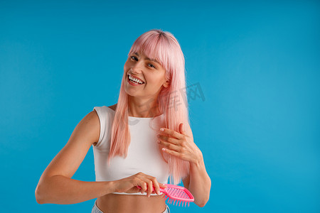 开朗的女人用梳子梳理光滑的天然粉红色长发，同时在蓝色工作室背景中摆出孤立的姿势