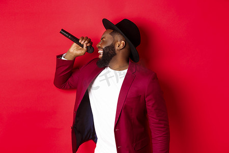 男唱歌摄影照片_热情的黑人男歌手在红色背景下表演，对着麦克风唱歌，穿着派对服装，站在红色背景上