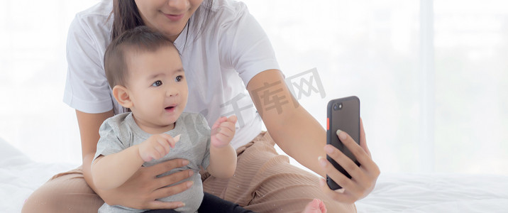 年轻的亚洲母亲和小女婴或新生儿在卧室的床上用智能手机自拍，幸福的母女在家里使用电话视频通话，两个人，家庭和沟通概念。