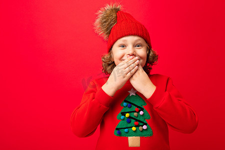冬天红墙摄影照片_穿着红色圣诞毛衣的酷少年在红墙、温暖的帽子和带圣诞树的毛衣的背景下闲逛