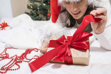 打开的红色礼盒摄影照片_戴着圣诞老人帽子的快乐的年轻黑发女人打开一个躺在白色床上的圣诞礼盒。