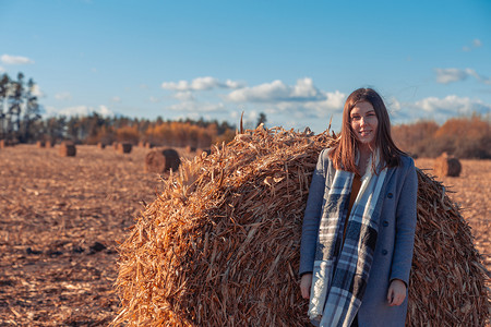 一个穿着灰色外套的欧洲女孩站在田野里，附近有一个更大的草捆，蓝天映衬着干草。