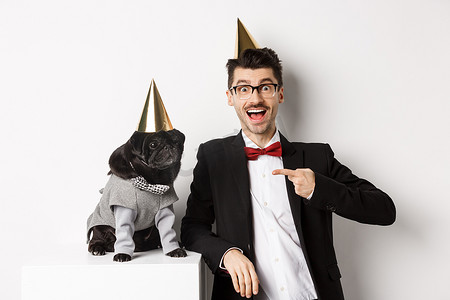 快乐的男人和可爱的黑色哈巴狗穿着派对锥和西装，狗主人庆祝宠物生日，站在白色背景上