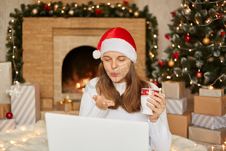 圣诞节在线祝贺，戴着圣诞老人帽子和白色毛衣的女孩与使用笔记本电脑进行视频通话的人交谈，发送飞吻，拿着茶杯，房间装饰得喜庆。