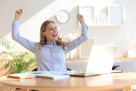 快乐的女企业家坐在办公桌前阅读好消息，举起双手表达喜悦。