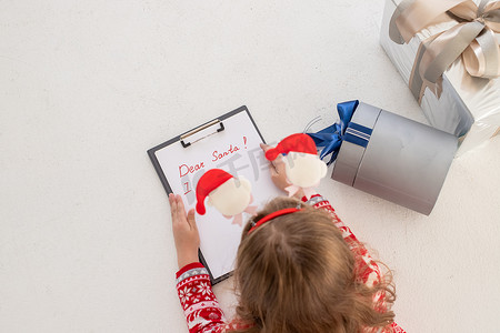 拿笔写字背景摄影照片_亲爱的圣诞老人信，圣诞贺卡。穿着红色毛衣的女孩，拿着毡笔，在白色背景的白纸上写字。童年梦想着礼物。圣诞节和新年概念。冬季假期