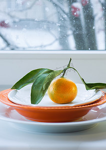 有窗背景摄影照片_雪窗背景下盘子上有树枝和叶子的橘子