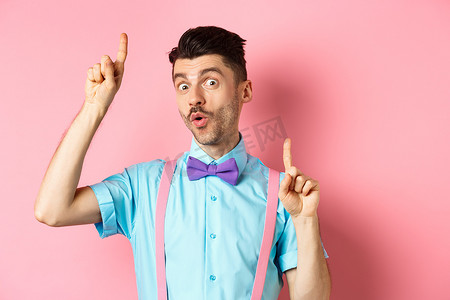 穿着领结和吊带跳舞的快乐有趣的男人，手指向上，看起来很乐观，站在粉红色的背景上