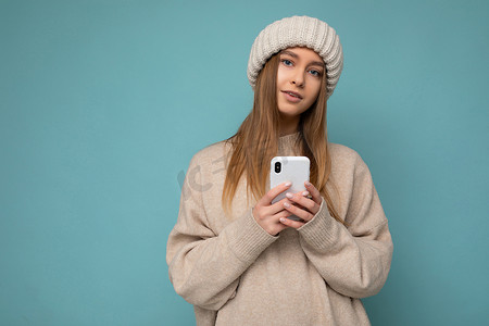 照片中，美丽、积极的年轻金发女子穿着时尚的米色温暖毛衣，头戴蓝色背景的针织冬季温暖米色帽子，手里拿着空的空间，并使用手机短信看着相机
