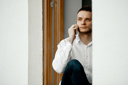 开窗户的人摄影照片_一名男子坐在开着的窗户上，看着街道，打电话。