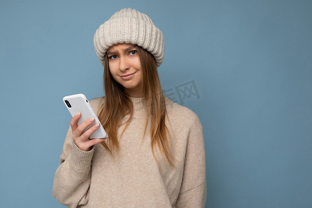 照片中，美丽的、心烦意乱的年轻金发女子穿着时尚的米色温暖毛衣，戴着蓝色背景上隔离的针织冬季温暖米色帽子，手里拿着空的空间，使用手机短信看着相机并提出问题