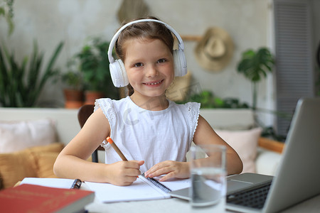 戴着耳机的微笑小女孩在家中使用笔记本电脑在线手写学习，戴着耳机的可爱快乐小孩子在电脑上上互联网网络课程或课程。