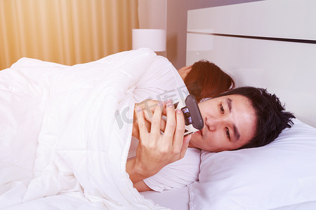玩手机的男人摄影照片_男人在床上玩手机，而他的妻子就睡在他旁边