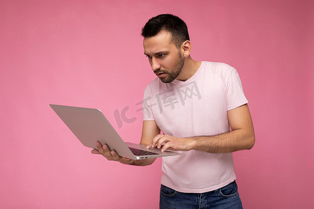 努力学习背景摄影照片_帅气的深色男子拿着笔记本电脑，在键盘上打字，看着穿着 T 恤的上网本显示器，背景是孤立的粉红色