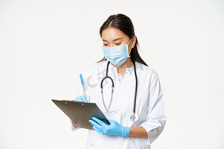 写下摄影照片_亚洲女医生在剪贴板上写下患者信息、写处方或诊断、戴着橡胶手套、白色背景的医用面罩
