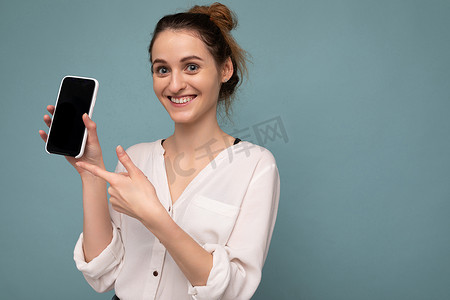 美丽积极的年轻金发卷发女子穿着休闲白衬衫，孤立在蓝色背景墙上，拿着电话，展示智能手机，上面有空的屏幕，用于模型查看相机并用手指指着设备