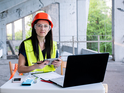 戴着防护头盔和护目镜的女建筑师在建筑工地新项目的个人电脑上工作
