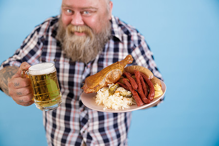 蓝色背景的肥胖男子，专注于手里拿着一盘油腻食物和啤酒的手