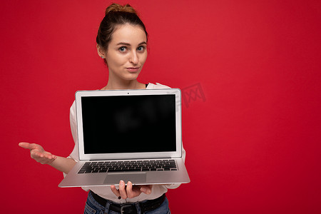 美丽微笑的快乐年轻女子的特写肖像，她拿着电脑笔记本电脑，看着相机，穿着休闲智能衣服，与红墙背景隔离，有疑问