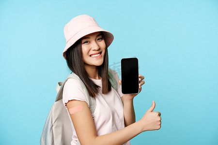 微笑的亚洲游客展示手臂上贴有covid-19疫苗接种补丁，在智能手机应用程序上展示冠状病毒疫苗国际证书，竖起大拇指