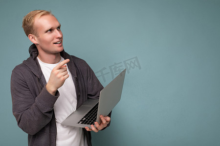 侧面照片中，英俊体贴的金发男子拿着电脑笔记本电脑，穿着白色 T 恤和灰色毛衣，抬头看着侧面，在蓝色背景上孤立地思考。
