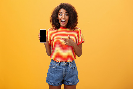 身穿条纹T恤、迷人的时尚苗条非洲裔美国卷发女性拿着令人惊叹的智能手机，指着橙色墙壁上的设备屏幕，惊讶得下巴都掉了下来