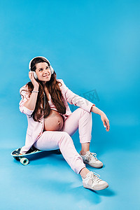 一位身穿粉色西装的孕妇戴着耳机坐在滑板上，听蓝色背景的音乐