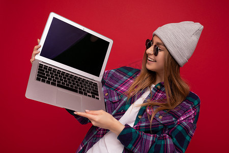 肖像照片中，美丽、滑稽、微笑的金发年轻女子拿着电脑笔记本电脑，显示器屏幕空空如也，戴着太阳镜帽子和彩色衬衫，看着上网本显示屏和红墙背景上隔离的键盘