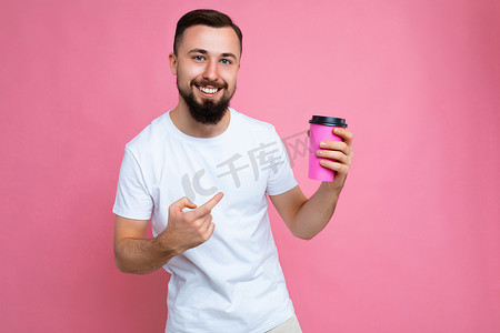照片中，英俊、快乐、黑发、胡子拉碴的年轻男子，留着胡子，穿着白色 T 恤，孤立在粉红色背景墙上，手里拿着纸咖啡杯，喝着酒，看着相机，用手指指着饮料